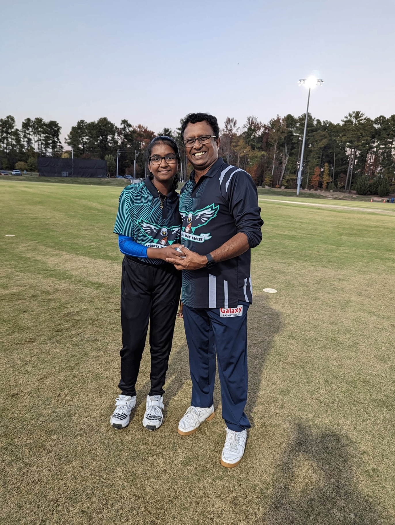 Head Coach Sudhir Nair with an academy player after a match, holding a cricket ball on the field